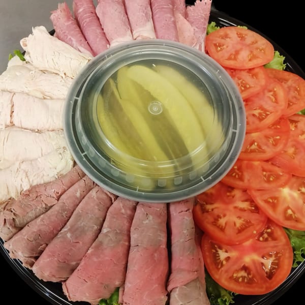 Deli platter with sliced turkey, ham, roast beef, tomatoes, lettuce, and a container of pickles.