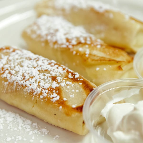 Three golden-brown blintzes dusted with powdered sugar, served with a side of sour cream.