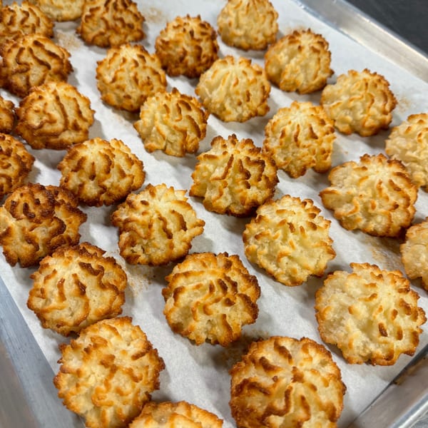 Many golden-brown coconut macaroons with textured tops are arranged on a white baking sheet.