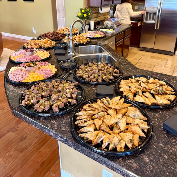 A kitchen counter laden with catering trays of ham and cheese cubes, skewers, and pastries.