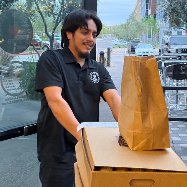 New York Bagels `N Bialys employee in black polo shirt with logo, pushing a stack of bagel boxes.