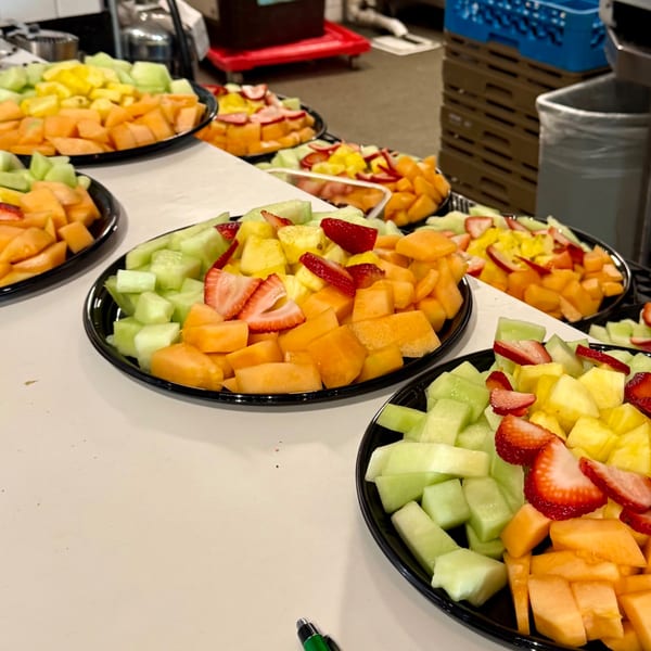 Multiple black platters filled with colorful, diced fresh fruit, including cantaloupe, honeydew, pineapple, and strawberries.