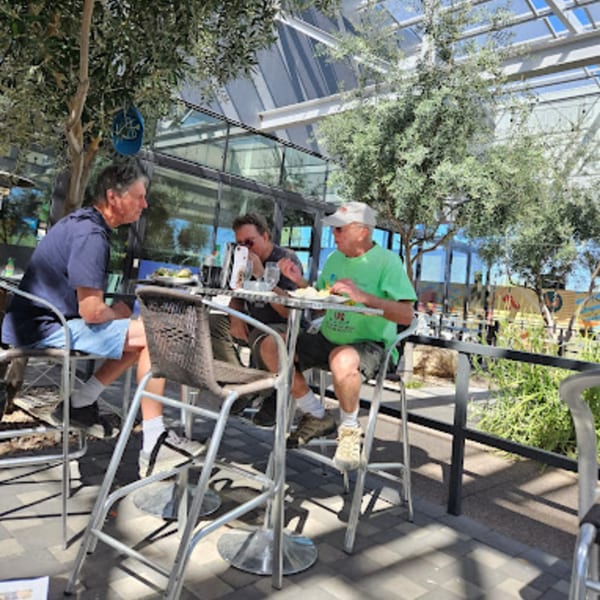 Three men enjoying bagels and drinks at an outdoor patio table under trees and a glass roof.