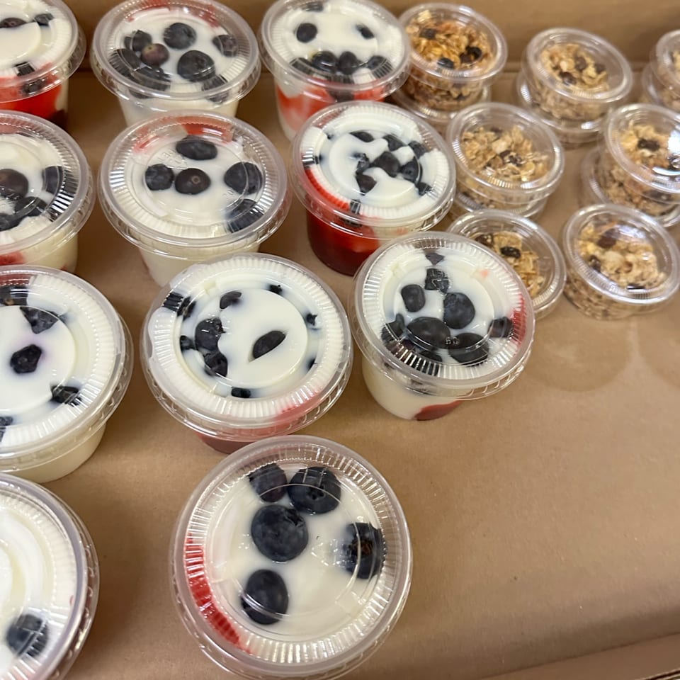Numerous clear plastic cups filled with yogurt and blueberries, and some with granola, in a box.