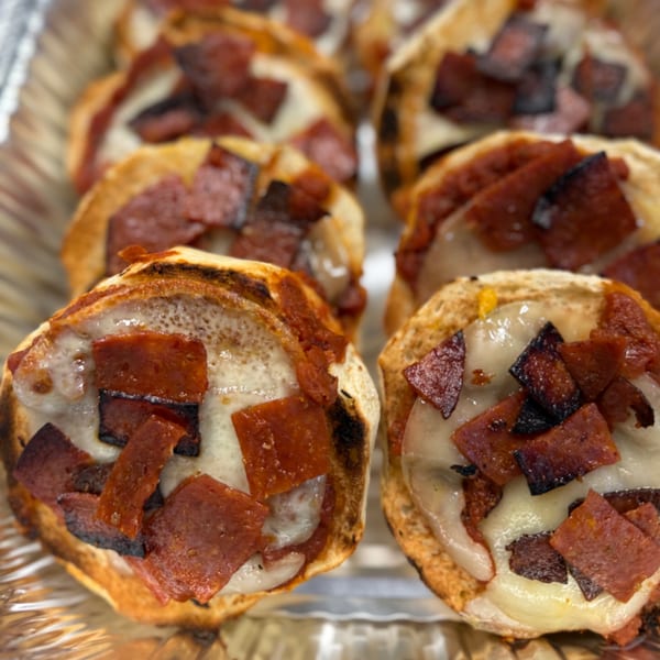 Close-up of several mini pizza bagels with melted cheese and pepperoni in a foil tray.