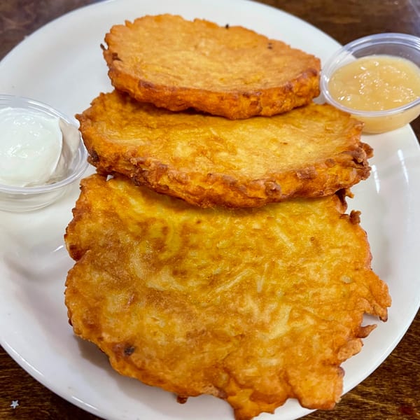 Three golden-brown latkes on a white plate with small cups of sour cream and applesauce.