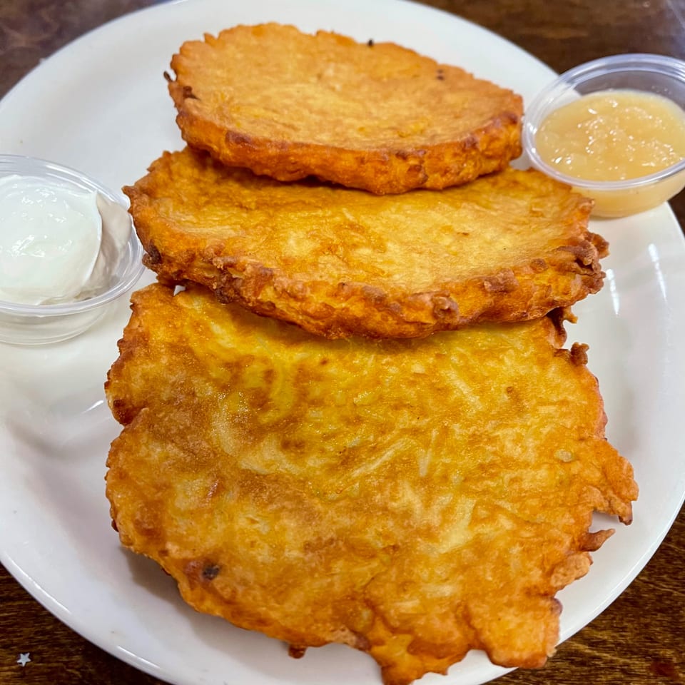 Three golden-brown latkes on a white plate with small cups of sour cream and applesauce.