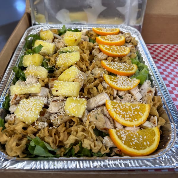 Chicken salad with pineapple, orange slices, sesame seeds, and crispy noodles in a foil tray.