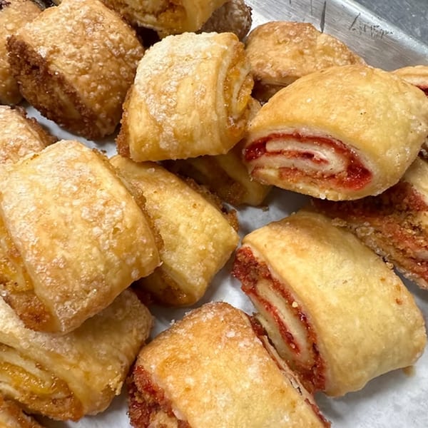 Close-up of golden-brown rugelach pastries, some with visible red fruit filling, on a white surface.